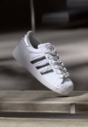 Biały sneaker Adidas z czarnymi paskami, grubymi sznurówkami i teksturowanym noskiem, spoczywający na szarej powierzchni w miękkim świetle.