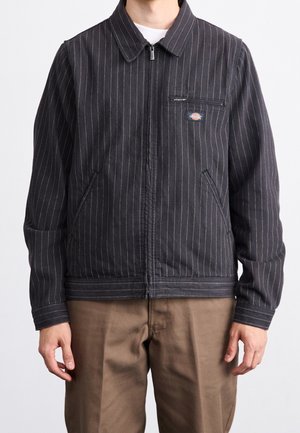 Mann trägt dunkelgraue Nadelstreifen-Zip-Up-Jacke mit Reißverschlusstasche auf der Brust und braune Hose, steht vor weißem Hintergrund.