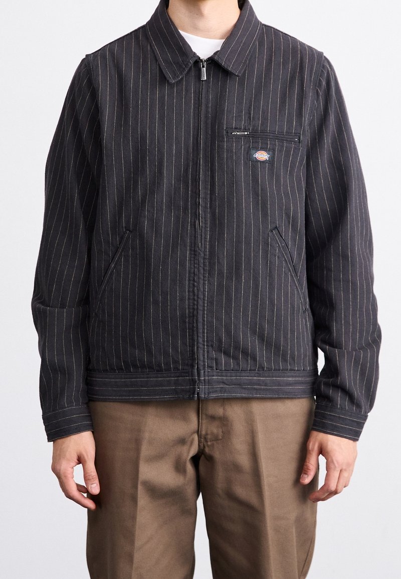 Homme portant une veste zippée gris foncé à fines rayures avec poche zippée sur la poitrine et pantalon marron, debout sur fond blanc.