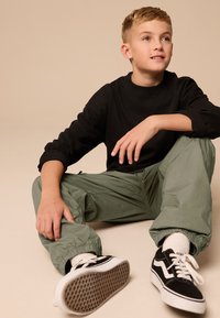 Chemise noire à manches longues, pantalon cargo vert avec poignets élastiques, et baskets Vans noir et blanc. Position assise avec les mains posées sur les jambes.