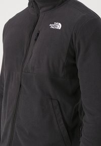 Zbliżenie na osobę w czarnej polarowej kurtce z kieszeniami na zamek i białym wyszywanym logo The North Face na klatce piersiowej.