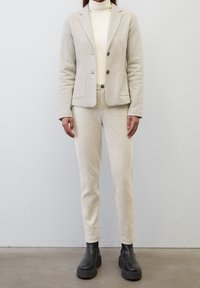 Béžový texturovaný blazer se dvěma knoflíky, odpovídající úzké kalhoty a černé robustní boty. Obsahuje přední kapsy a krémový top s vysokým límcem.