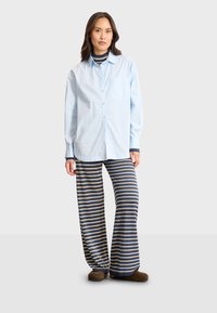 Camicia azzurra chiara con bottoni e taschino sul petto, indossata sopra un pantalone da pigiama a righe in blu navy, beige e bianco. Tessuto morbido, vestibilità rilassata.
