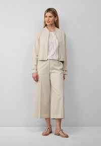 Beige cropped bomberjack, wit gepatternde top, wijde beige broek en sandalen met strikaccenten. Gladde stof, minimalistisch ontwerp.