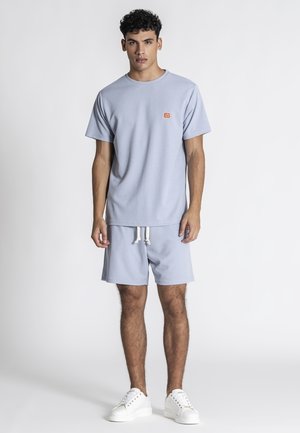 Junger Mann steht, trägt hellblaues Kurzarmhemd und passende Shorts mit weißen Turnschuhen vor einfachem weißem Hintergrund.