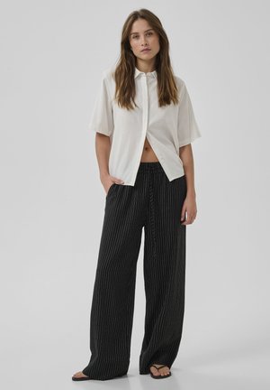 Nő áll, kezét a zsebében tartva, laza, fehér, rövid ujjú gombos ingben, fekete, csíkos, bő szárú nadrágban és szandálban.