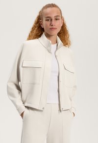 Helle beige Jacke aus weichem Stoff, mit Stehkragen, zwei Brusttaschen und Druckknopfverschluss. Glattes, minimalistisches Design.