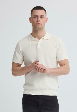 Man met kort bruin haar die een crème polo met korte mouwen en donkere broek draagt, staand met losjes gevouwen handen.