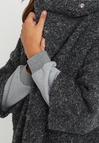 Dunkelgrauer, strukturierter Oversized-Mantel mit hohem Kragen, der mit Druckknöpfen geschlossen wird, über einem hellgrauen Sweatshirt getragen.