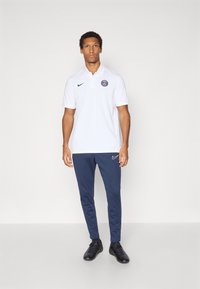 Hvid polo t-shirt med knappet krave og logo, parret med navy tapered sweatpants med et sidelogo og sorte sportsko.