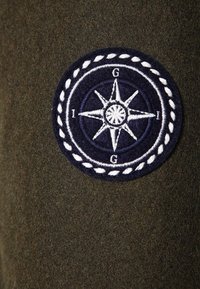 Una tela de color verde oscuro cuenta con un parche bordado circular en azul marino con un diseño de compás blanco y un borde parecido a una cuerda, que incluye las iniciales "G" e "I."
