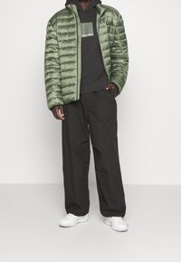 Man draagt een groene donzen jas over een zwarte Calvin Klein hoodie, zwarte losse broek en witte sneakers, staand met één hand in zijn zak.