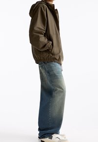 Chaqueta ligera verde oliva con cremallera, capucha y dobladillo elástico, combinada con vaqueros azules descoloridos y zapatillas blancas.