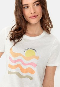 Witte katoenen T-shirt met een golvend grafisch ontwerp met oranje, roze, gele en groene lijnen, onder een cirkelvormige gele zon. De tekst luidt "waterloop."