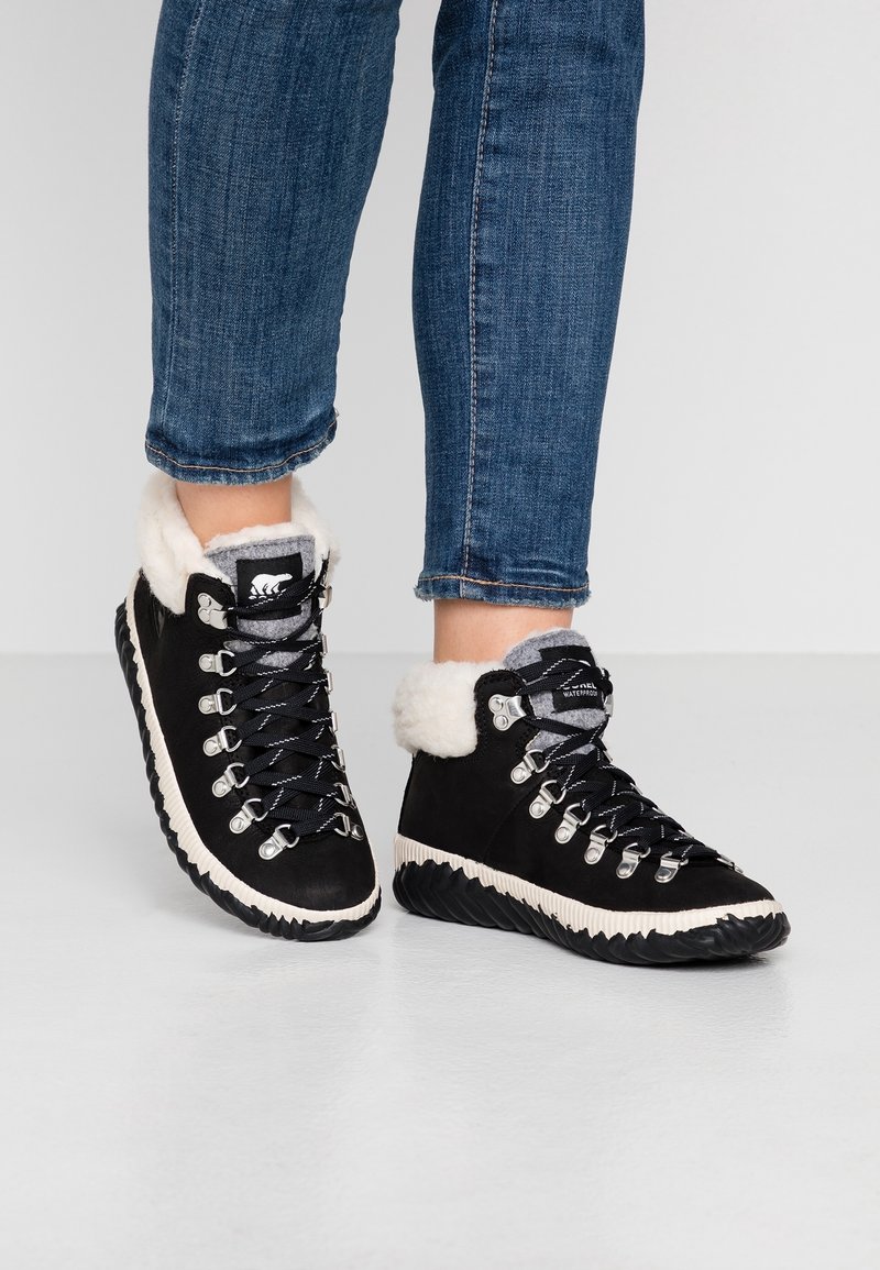 Personne portant des bottes d'hiver montantes noires avec une doublure en fourrure blanche et des semelles épaisses noires, associées à un jean en denim bleu.