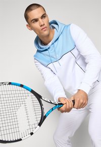 Sudadera blanca con capucha y panel superior en azul claro, con un logo. El modelo sostiene una raqueta de tenis negra y azul, de pie contra un fondo neutro.