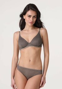 Donna con lunghi capelli scuri che indossa un reggiseno e delle mutandine in pizzo taupe coordinati, in piedi contro uno sfondo chiaro e neutro.