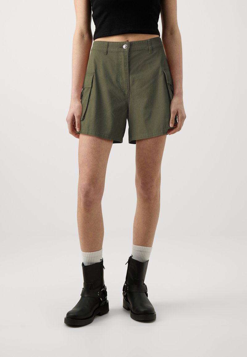Personne portant un short cargo vert olive, un crop top noir, des chaussettes blanches et des bottines noires, debout devant un fond clair uni.