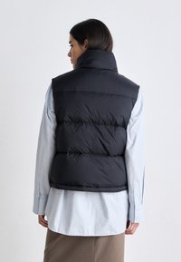 Svart puffervest med høy krage, quiltet tekstur og runde armhull, båret over en lyseblå skjorte og brun skjørt.