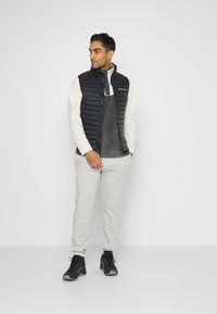 Gilet imbottito nero sopra un top in pile grigio, abbinato a pantaloni della tuta grigio chiaro e scarpe da ginnastica nere. Design semplice, senza motivi visibili.