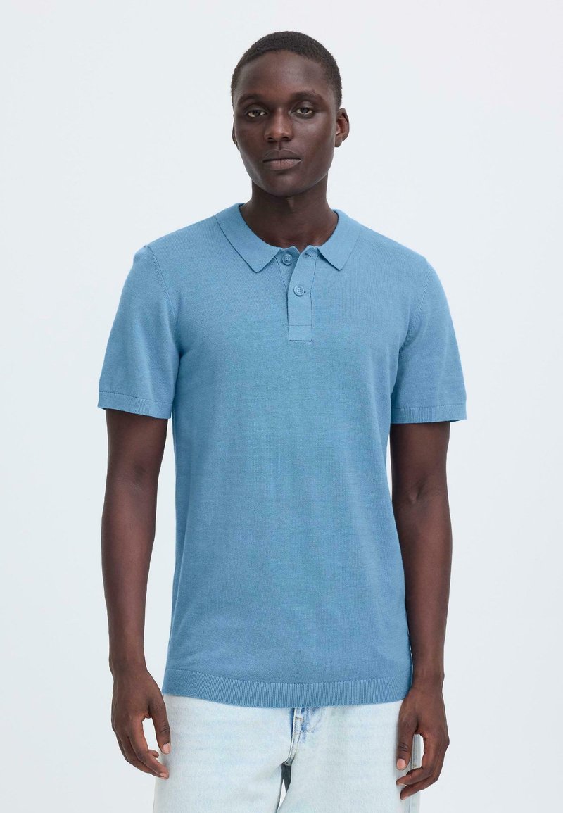 Polo bleu clair tricoté avec col, manches courtes et patte de boutonnage ; présente une texture lisse et un ourlet côtelé.