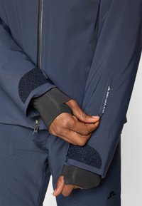 Casaco impermeável azul-marinho com punhos em velcro, tecido elástico, bolso com fecho éclair e detalhes cinzentos contrastantes no fecho e na manga.