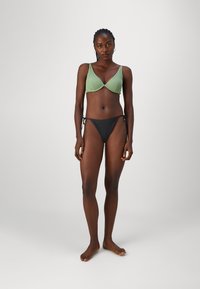 Zeleno rebrast bikini zgornji del z tankimi naramnicami in globokim izrezom, v kombinaciji s črnimi bikini spodnjimi deli z vezanjem na straneh. Gladka tekstura, minimalističen dizajn.