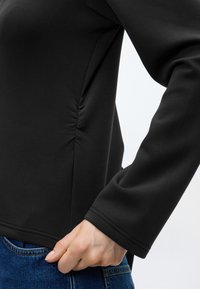 Personne portant un haut noir à manches longues avec un détail froncé sur le côté, ajustant l'ourlet près de la poche d'un jean bleu.