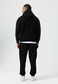 Zwarte hoodie met een grote capuchon en geribbelde boorden, gecombineerd met bijpassende zwarte joggers met een achterzak. Model draagt witte sneakers.