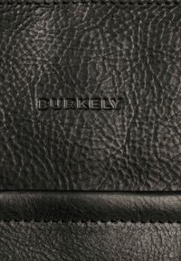 Material de cuero negro con una superficie texturizada, que presenta un logo "BURKELY" en relieve. Incluye detalles de costura a lo largo del borde.