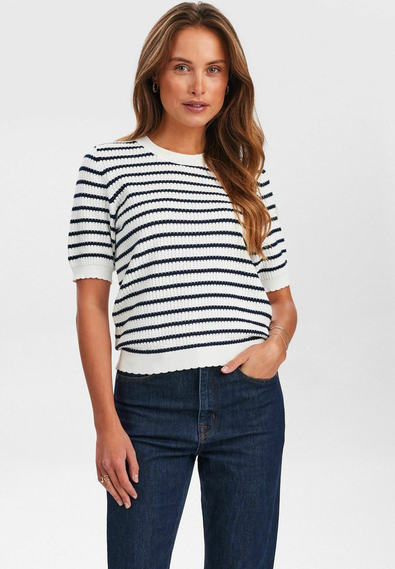 Balts un tumši jūras zils svītrots īsām piedurknēm adīts džemperis ar noapaļotu kakla izgriezumu un viļņotu malu. Valkāts kopā ar augsta vidukļa tumšas krāsas džinsiem.