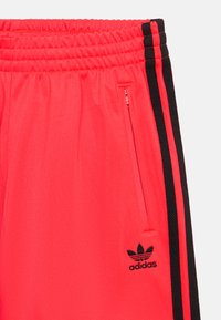 Raudonos sportinės kelnės su elastinga juosta, šoniniu užtrauktuko kišeniu ir juodomis šoninėmis juostelėmis. Apatinėje kojoje yra juodas "adidas" logotipas.