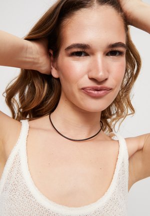 Collier ras du cou noir, fin et arrondi, porté avec un débardeur texturé blanc, sur un fond neutre.