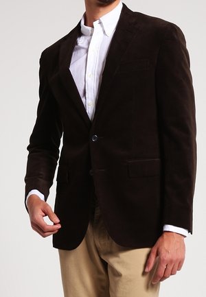 Uomo che indossa un blazer marrone scuro sopra una camicia bianca con bottoni e pantaloni beige, in piedi davanti a uno sfondo semplice.
