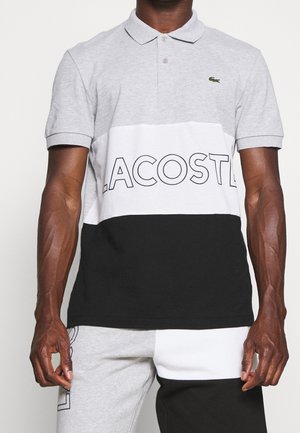 Mand iført en tre-farvet polo med teksten "LACOSTE" og krokodillelogo, kombineret med matchende sorte, hvide og grå shorts.