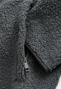 Tessuto in pile grigio con una finitura strutturata e soffice. Presenta un'apertura con zip, che mostra un tessuto interno liscio.