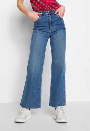 Persona con vaqueros azules de tiro alto y pernera ancha, camiseta roja de rayas, zapatillas blancas gruesas, manos en los bolsillos frontales, de pie frente a un fondo blanco.