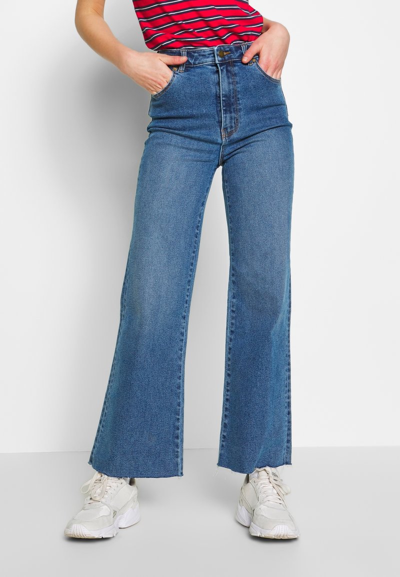 Personne portant un jean bleu taille haute à jambes larges, un haut rouge à rayures, des baskets blanches épaisses, les mains dans les poches avant, debout sur fond blanc.
