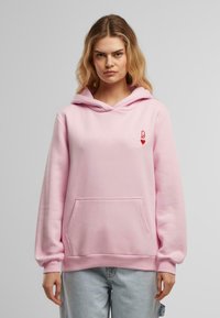 Haut rose en molleton, avec une poche avant, capuche à cordon et un graphique en forme de cœur rouge sur la poitrine. Bordures côtelées aux poignets et à l'ourlet.
