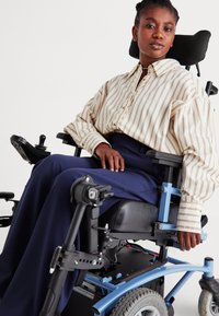 Elektrische rolstoel met een zwarte zitting, blauw frame en grijze wielen. Gebruiker in een gestreept crèmekleurig en bruin overhemd met losse mouwen.