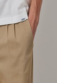 T-shirt en coton blanc avec une étiquette portant la mention "studio" et pantalon beige plissé présentant une texture lisse et une coupe ajustée.