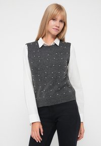 Femeie îmbrăcată cu un veston gri fără mâneci, decorat cu mici perle albe, peste o cămașă albă cu guler și pantaloni negri, stând în fața unui fundal alb.