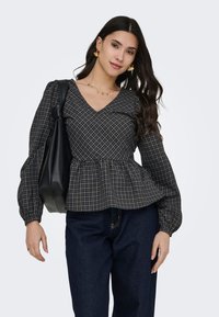 Femme aux longs cheveux foncés portant des boucles d'oreilles et un collier en or, un chemisier noir à carreaux avec col en V et manches bouffantes, un jean foncé, et un sac à bandoulière noir.