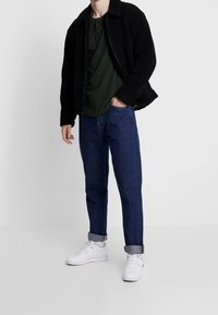 Chaqueta negra texturizada sobre una camiseta henley verde, combinada con jeans azules y zapatillas blancas, mostrando un look casual y en capas.