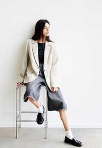 Oversized beiger Blazer über einem schwarzen Oberteil, kombiniert mit grauen Jeans-Culottes, weißen Socken und schwarzen Loafers, sitzt auf einem metallischen Hocker.