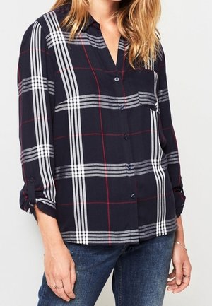 Femme portant une chemise boutonnée à manches longues bleu marine avec un motif à carreaux blancs et rouges, assortie à un jean bleu.