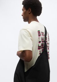 Κρεμ κοντομάνικη μπλούζα με αφηρημένο πράσινο και ροζ κείμενο στην πλάτη. Φοριέται μαύρη τσάντα ώμου με λεία υφή.