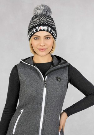 Black and white knit beanie with gray pom-pom, featuring a Ski Austria logo. A fitted gray vest with zip pockets and a hood is worn underneath.