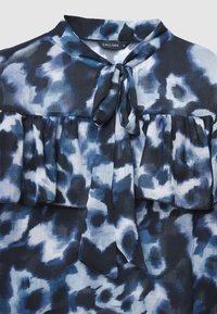 Dunkelblaue Bluse mit Kragen, Bindungsdetail und Rüschendetails. Sie hat ein abstraktes blau-weißes Muster auf leichtem Stoff.