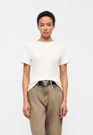 Kobieta ubrana w prostą białą koszulkę z krótkim rękawem, wpuszczoną w beżowe spodnie z czarnym paskiem z ozdobną, srebrną klamrą w kształcie serca.